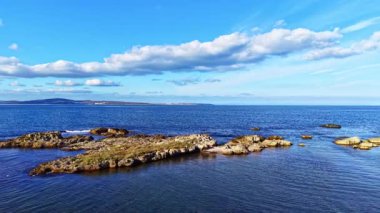 Sakin bir sahil sahnesinde, kabarık bulutlarla dolu canlı bir gökyüzü altında kayalık oluşumlarla karşılaşan bir su birikintisi görülüyor. Ortam huzur ve doğal güzelliği yansıtıyor..
