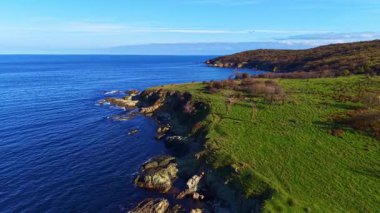 Dalgalar kayalık kıyılarda yavaşça dolaşırken parlak sabah ışığı suda dans eder. Bereketli yeşil tepeler ışıl ışıl denizle buluşmak için yamaçlara iniyor, dingin ve pitoresk bir manzara yaratıyor..