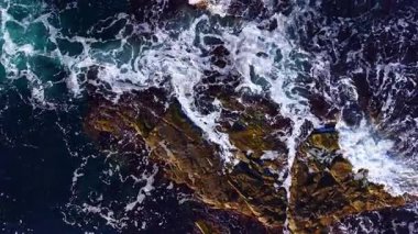 Güneş batarken dalgalar engebeli kayalarla çarpışır ve havaya köpük püskürtür. Sahne, okyanusun vahşi güzelliğini ve ham gücünü solgun ışığın arkaplanına yansıtıyor..