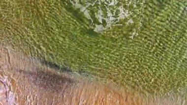 Berrak sular altın kumun üzerinde yavaşça dans eder. Güneş yukarıdan aydınlık bir şekilde parlarken yatıştırıcı desenler yaratır. Ziyaretçileri deniz kenarında huzurlu bir öğleden sonranın tadını çıkarmaya davet eder..