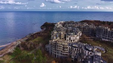 Sakin bir sahil manzarasında, su kenarına yakın çökmüş binalar doğanın kucağına sarılmış. Gökyüzü bulutlarla kaplı, bu unutulmuş yerde unutulmaz bir güzelliği yansıtıyor..