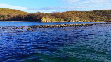 Yumuşak dalgalar, parlak bir gökyüzünün altında yeşil tepelerle çevrili kayalık bir sahile nazikçe çarpar. Huzurlu sahne, barışçıl bir yansıma ve doğanın güzelliğini takdir etmeye davet ediyor..