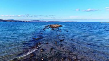 Sakin denize doğru uzanan kayalık bir oluşum, gün ortası güneşini yansıtan hafif dalgalarla çevrilidir. Manzara, berrak mavi bir gökyüzünün altında huzurlu bir atmosfer yaratan uzak tepeleri barındırıyor..