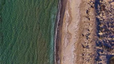 Kristal berrak sular, güneşli bir sahile doğru yavaşça dalgalanır. Kumlu sahil, sıcak öğle güneşinin altında yumuşak dalgalarla buluşur. Huzurlu ve huzurlu bir manzara yaratır..