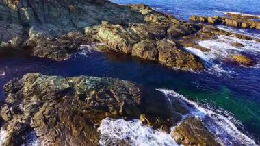 Güneş ışığı kıyı manzarasını aydınlatırken dalgalar yavaşça engebeli kayalara çarpıyor. Turkuaz su, güzel bir günde sakin bir atmosfer yaratan titreşimli gökyüzünü yansıtır..