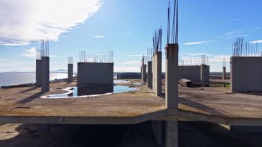 Beton sütunlar, suyun yakınındaki tamamlanmamış bir inşaat sahasında parlak mavi gökyüzünü ve dağınık bulutları yansıtıyor. Bir su birikintisi olay yerine huzur katar..