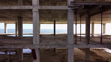 Terk edilmiş bir bina kısmen tamamlanmış, ham beton çerçevesi elementlere maruz kalmış. Uzakta deniz ışıldıyor, ıssız uzayda çarpıcı bir kontrast oluşturuyor..