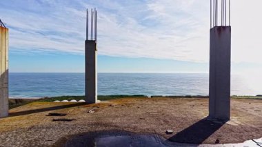 Güneş ışığı, bitmemiş beton sütunların yer aldığı bir inşaat sahasındaki sakin okyanus suyundan yansıyor. Huzurlu manzara doğa ve sanayi arasında bir zıtlık yaratır..
