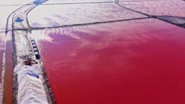 Geniş tuz düzlükleri açık bir gökyüzü altında pembe ve beyaz renkleri paylaşır. Yakın makineler ve tuz yığınları doğanın büyüleyici sanatsal sahnesine eklenir.