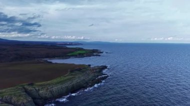 Engebeli uçurumlar ve canlı yeşil alanlar denizin derin mavi sularıyla tezat oluşturuyor. Bulutlar kasvetli bir gökyüzünde sürükleniyor ve alacakaranlıkta sakin bir manzara yakalıyor.