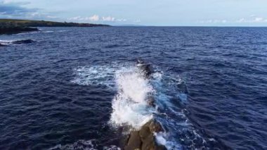 Okyanus dalgaları dans eder ve kayalık oluşumlara çarparak parlak bir gökyüzünün altında beyaz köpükler saçar. Huzurlu sahne engebeli bir kıyı şeridi boyunca uzanıyor, bir huzur hissi uyandırıyor..