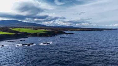 Yeşil alanlar kıyı şeridi boyunca uzanırken dalgalar yavaşça koyu kayalara çarpar. Görkemli dağlar bulutlarla dolu dramatik bir gökyüzünün altında yükseliyor..