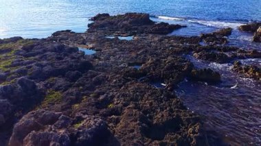 Sabahın erken saatlerinde engebeli bir kıyı şeridinde parıldayan ışık karanlık kayaları ve titreyen gel-git havuzlarını ortaya çıkarıyor. Sakin dalgalar toprağı kucaklıyor, yansıma için mükemmel huzurlu bir atmosfer yaratıyor..