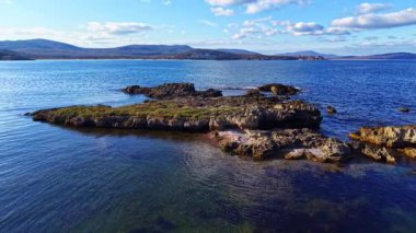 Berrak sular, sabah ışığını kıyıdan çıkan kayalık oluşumlar olarak yansıtır. Ufuk, parlak, mavi bir gökyüzünün altında tüylü bulutlarla kaplı uzak tepeleri ortaya çıkarıyor..