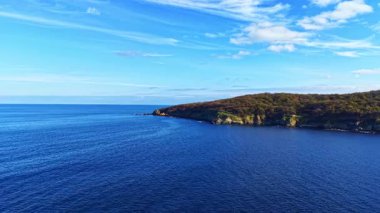 Canlı mavi sular, parlak gökyüzünde kabarık bulutların sürüklenmesi gibi yeşil tepelere nazikçe sarılır. Bu sakin sahne huzurlu bir öğleden sonra doğanın güzelliğini gözler önüne seriyor..