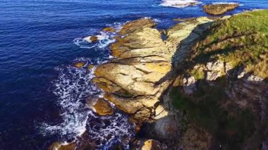 Parlak güneş ışığı taşlara çarpan dalgalar gibi kayalık kıyıları aydınlatır. Derin mavi okyanus kayalık dokularla çelişiyor. Huzurlu ama dinamik bir manzara yaratıyor..