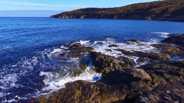Engebeli kayalar canlı mavi okyanustan yükselirken yumuşak dalgalar üzerlerini kaplıyor ve sakin bir atmosfer yaratıyor. Güneş, sakin sahnenin üzerine doğanın güzelliğini arttıran sıcak bir parıltı saçar..