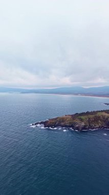Yumuşak sabah ışığı, dalgaların engebeli bir kıyı şeridine ulaştığı sakin bir kıyı sahnesini gözler önüne seriyor. Yukarda kayan tepeler ve bulutlu gökyüzü ile çevrili..