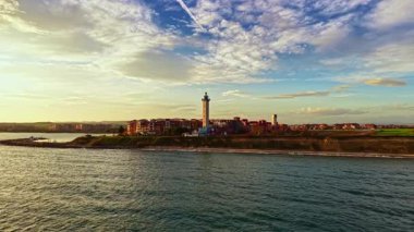 Altın güneş ışığı, deniz fenerinin durduğu sakin suya yansıyor. Arka planda kayan tepelerle, deniz kenarındaki tuhaf bir köyü gözlüyor. Kıyı yaşamının özünü yakalıyor..