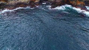 Berrak mavi sular, dalgaların kayalık kıyı şeridinde ritmik bir şekilde kırılmasıyla güneş ışığını yansıtıyor ve sahil kenarında huzur ve sükunet hissi yaratıyor..