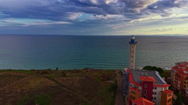 Sahil şeridi yakınlarındaki canlı binaların yanında bir deniz feneri kulesi bulutların toplanmasıyla sakin suların üzerine gölgeler düşürüp huzurlu bir günbatımının sinyallerini veriyor. Hava sükunetle dolu..