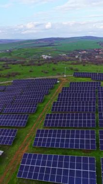 Güneş panellerinin alanları yeşil arazide düzenli olarak düzenlenir. Manzara, gündüz vakti parlak bir gökyüzü altındaki tepeleri ve uzak binaları içerir..