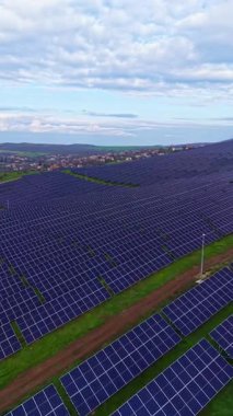 Güneş panelleriyle kaplı geniş bir alan yeşil bir alan boyunca uzanıyor. Paneller düzgün bir şekilde hizalanır, parçalı bulutlu bir gökyüzünün altında ışığı yansıtır. Yakın yapılar uzaklıktan görülebilir..