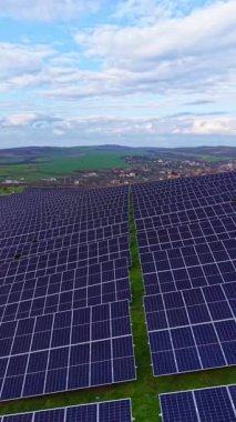 Güneş panelleri kırsal bir alanda kırsal alana yayılıyor. Gökyüzünde bulutlar var ve arka planda tepeler görülebilir. Yakınlarda küçük bir kasaba var..
