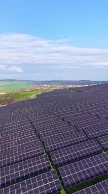 Birçok güneş paneli bir yamacı doldurur, üzerinde açık mavi bir gökyüzü ve arka planda yeşil alanlar vardır. Bu kurulum yenilenebilir enerji projesinin bir parçasıdır..