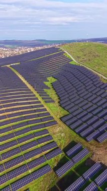 Güneş panelleri tepedeki büyük bir alanı kaplayarak güneş ışığını topluyor. Yeşil çimenler panelleri sarar ve manzara mavi gökyüzünün altında biraz bulutla birlikte uzanıyor..