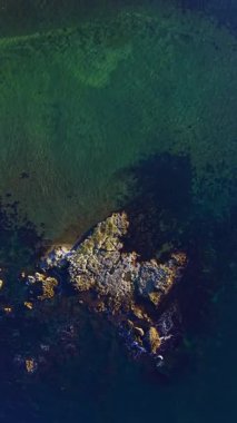 Kıyı şeridi boyunca uzanan berrak sulardan kayalar görünür deniz yosunu desenleri ve yüzeyden yansıyan güneş ışığı tarafından oluşturulan gölgelerle ortaya çıkar..