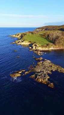 Yakınında yeşil bir tepe olan kayalık bir kıyı şeridi suya doğru uzanır. Deniz sakin ve berrak bir gökyüzü görünüyor. Bu sahne gündüz çekiliyor..