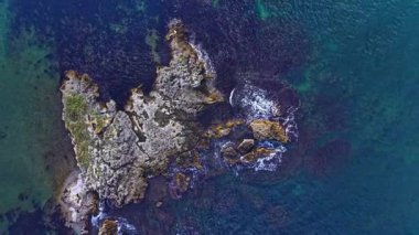 Pürüzlü ve kayalık oluşumları çevreleyen berrak mavi sularda parlak güneş ışığı parıldıyor. Yumuşak dalgalar yavaşça kıyılara vurur ve doğanın güzellikleriyle süslenmiş sakin bir sahil atmosferi yaratır..