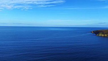 Sakin sular güneş ışığı altında parlar, ufukta gökyüzü ile pürüzsüz bir şekilde birleşir. Engebeli bir kıyı şeridi huzur ve uçsuz bucaksızlık hissi sunan sakin sahneyi özetliyor..