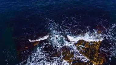 Güçlü dalgalar, deniz yüzeyinde güneş ışığı dans ederken karanlık ve pürüzlü kayalarla çarpışır, sahneyi turuncu ve mavi renklerle boyarken sahil kenarında sakin bir akşam boyunca.