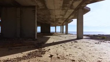 Güneş ışığı uzun zaman önce terk edilmiş bir binanın iskelet yapısının içinden akar. Çatlamış zeminde dramatik gölgeler oluştururken sakin deniz uzaklarda parıldar..