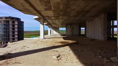 Uçsuz bucaksız, bitmemiş beton bir yapı terk edilmiş duruyor. Güneş ışığı boşluklardan akar, tozlu zeminde gölgeler oluştururken yeşil tepeler uzaklarda uzanır, derin düşüncelere daldırır.