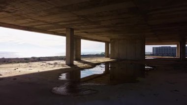 Güneş ışığı, terk edilmiş bir binanın iskelet kirişlerinden süzülerek uzun gölgeler bırakır. Su birikintileri gökyüzünü yansıtıyor, sessiz ve unutulmuş bir dünyayı ima ediyor..