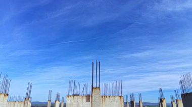 İşçiler beton sütunlar ve çıkıntılı demirle çevrili bir bina iskeleti inşa ediyorlar. Açık mavi gökyüzü bölgeyi aydınlatıyor ve gelişmeleri gösteriyor..