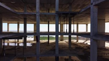 Tamamlanmamış bir yapı kıyıda tek başına duruyor, güneş ışığı kirişlerinden akıyor, sakin deniz ve ötesinde yeşil alanlar ile çelişiyor..