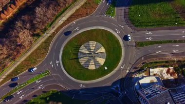 Araçlar yerde geometrik desenli bir döner kavşağın etrafında hareket ederler. Yollar farklı yönlere giden yollara bağlanır. Arka planda ağaçlarla çevrili yeşil çimenler var..