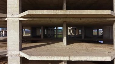 Geniş boş alanlar, yumuşak ışıkla aydınlatılmış beton bir binanın iskelet çerçevesini ortaya çıkarır. Doğa dışarıda gelişiyor, arka planda yeşil tepeler var, olası bir geleceği ima ediyor..