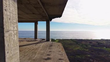 Terk edilmiş bir bina uçurumun kenarında duruyor, sakin okyanusu ve güneş doğmaya başladığında berrak gökyüzünü nefes kesici bir şekilde gösteriyor, altın ışıkla sahneyi aydınlatıyor..