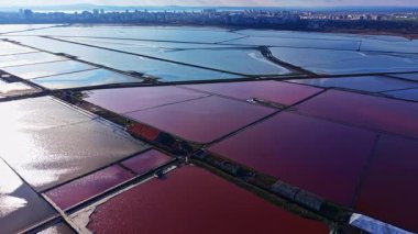 Canlı tuz düzlükleri güneş ışığında parıldıyor, güzel bir kırmızı ve mavi renk karışımı sergiliyor. Kentsel ufuk çizgisi arka planda duruyor, benzersiz manzarayı vurguluyor..