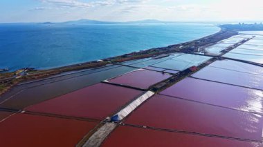 Durgun denizle tezat oluşturan çeşitli gölgelerde parlayan canlı tuz düzlükleri, berrak mavi gökyüzü ve uzak tepeler tarafından kucaklanan pitoresk bir manzara yaratıyor..