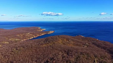 Geniş bir manzara, kahverengi karanın mavi denize çıktığı bir kıyı şeridini gösteriyor. Uzak tepeler açık bir gökyüzünün altında görülebilir. Öğlen oldu ve ufukta hiç bulut yok..