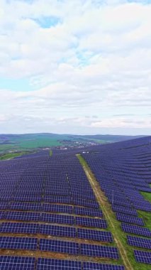 Güneş panelleri kırsal alanlarda geniş alanlara yayılıyor. Manzaranın arka planında tepeler ve yukarıda bulutlu bir gökyüzü var..