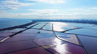 Renkli tuz düzlükleri kıyı boyunca güneş ışığında parıldarken, uzak bir ufuk çizgisi kentsel yaşamı işaret ediyor. Durgun su, gökyüzünün çarpıcı bir yansımasını yaratır..