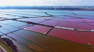 Geniş tuz düzlükleri açık gökyüzünü yansıtırken kırmızı, mavi ve kahverengimsi renkler sergiliyor, yakınlarda canlı bir kentsel ufuk çizgisi ile yan yana, doğa ve insan mimarisini gözler önüne seriyor..
