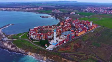 Binalar mavi bir su kütlesinin yanında eğimli bir kıyı şeridini kuşatır. Yapıların arkasında yeşil alanlar uzanıyor. Tepeler berrak bir gökyüzünün altında yükselir.